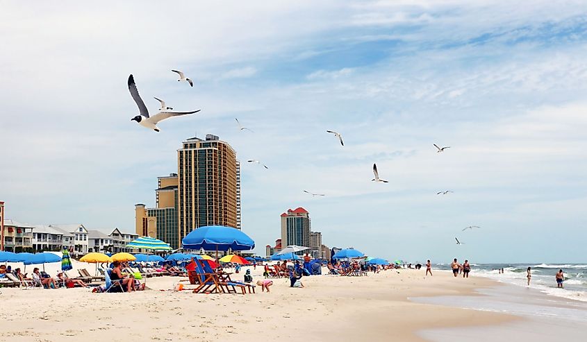 Marine landscape with beach line homes and hotels, beach umbrellas, people and flying birds on a foreground in Alabama Gulf Shores State park and beach, USA.