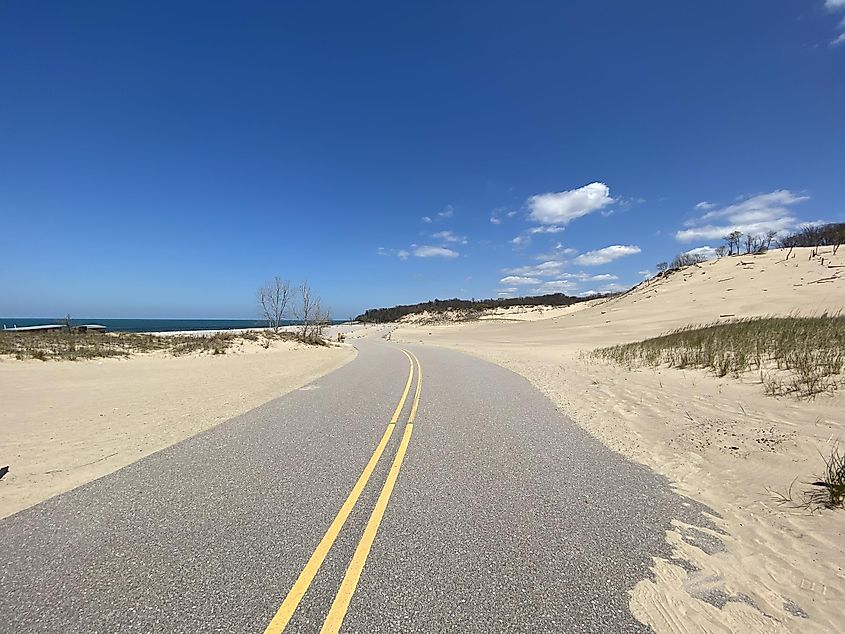 A sand swept road leads past a dune and toward a beachside parking lot