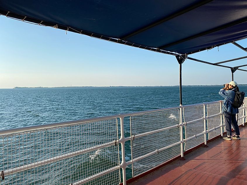 A woman standing aboard a ferry photographs an island in the distance.