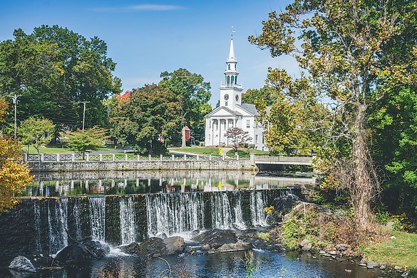 White church with a tall steeple overlooking a waterfall on the Wepawaug River in Milford, Connecticut, surrounded by trees and a small bridge.
