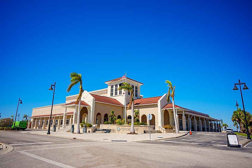 Charlotte Harbor Event and Conference Center in Punta Gorda, Florida, with a beige exterior, red-tiled roofs, palm trees swaying in the foreground, and a clear blue sky above.