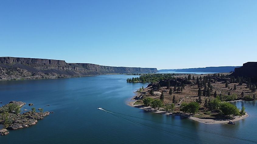 A serene lake scene with a small boat trailing waves across the blue water. Rocky cliffs and lush green trees border the lake under a clear, sunny sky.