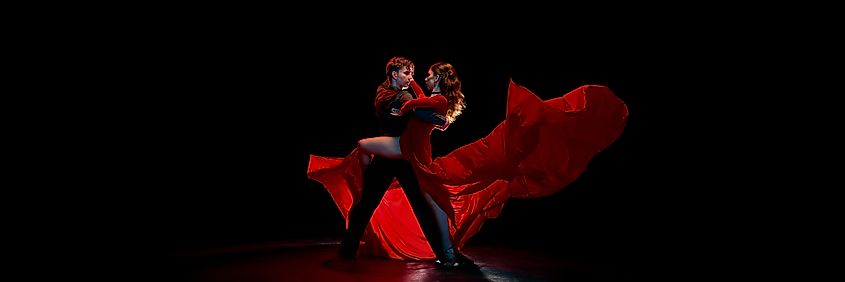 Dramatic dancers of Argentine tango passionately embrace on a dimly lit stage, with a flowing red dress highlighting the scene. Concept of intense dance chemistry within performance