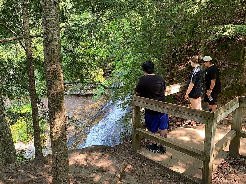A group of hikers stand atop a wooden observation deck beside a large forest waterfall.