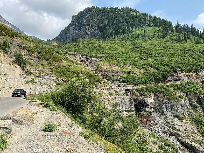 A smaller roadside turnout looking onto a tiered waterfall, arched bridge, and forested mountain peak. A classic Going-to-the-Sun Road scene.