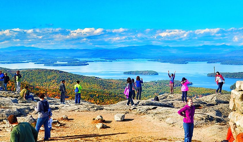 Mount Major, New Hampshire, tourists posing for photos at the summit. Lake Winnipesaukee and the Belknap Mountains in the background. Forest trees are changing color for the fall.