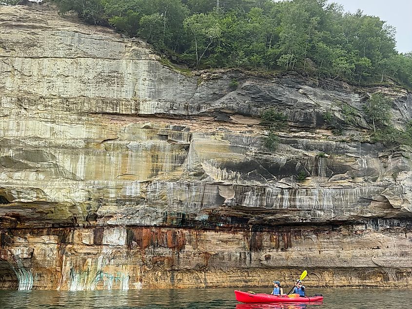 A tandem kayak passes a sheer cliff with colorful mineral stains.
