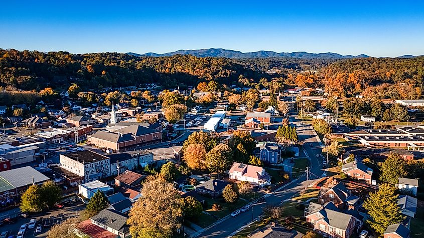Aerial view of a small town surrounded by lush autumn trees and distant mountains. Buildings and houses are scattered under a clear blue sky.