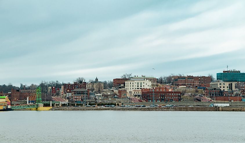 Cityscape of historic Alton, Illinois sprawled across the bluffs along the Mississippi River with the river in the foreground and a blue sky in the background.