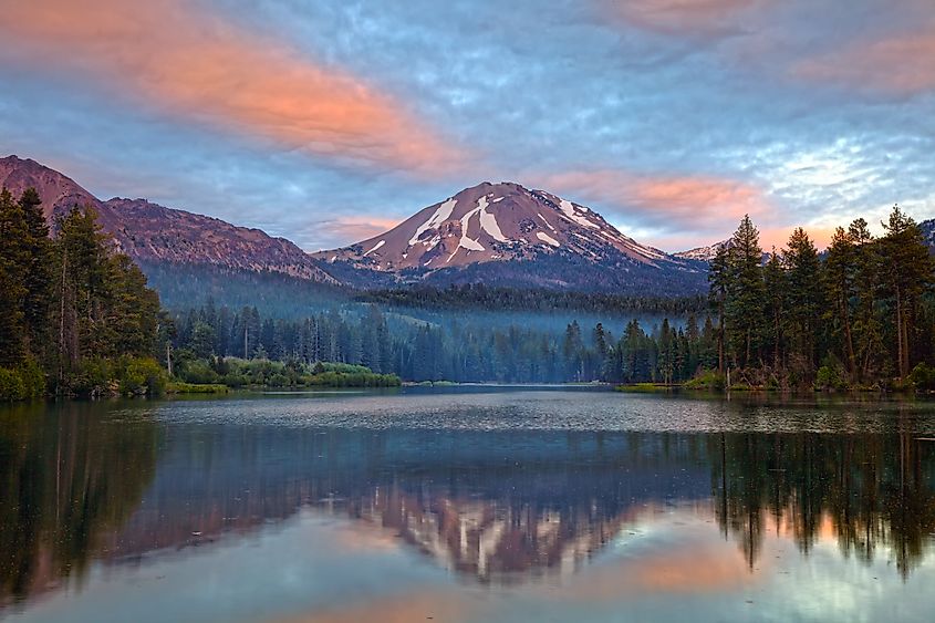 Serene lake with a mirror reflection of snow-capped mountain and pine trees at sunrise. Pink and blue clouds create a tranquil, peaceful atmosphere.