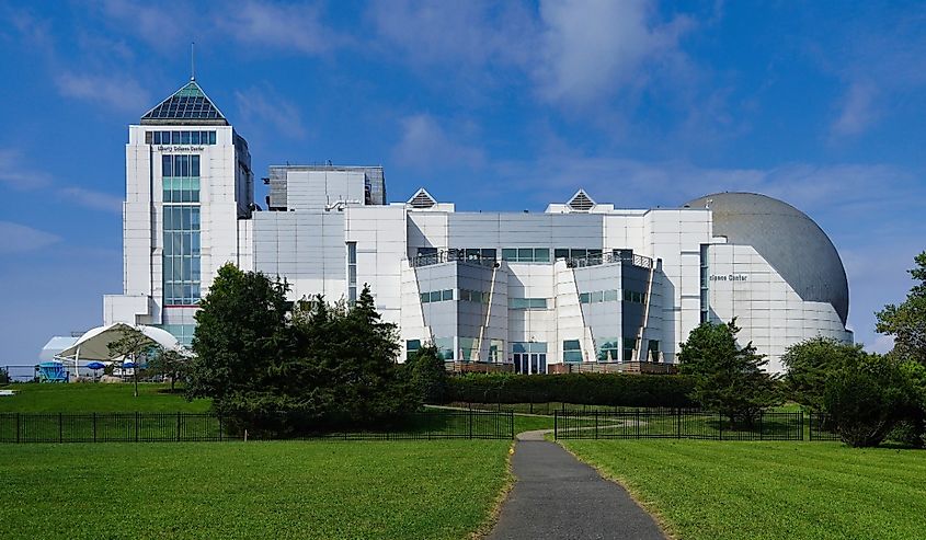 A modern science museum with a white, geometric facade and a large dome sits under a blue sky. A path leads through a green lawn to the entrance.