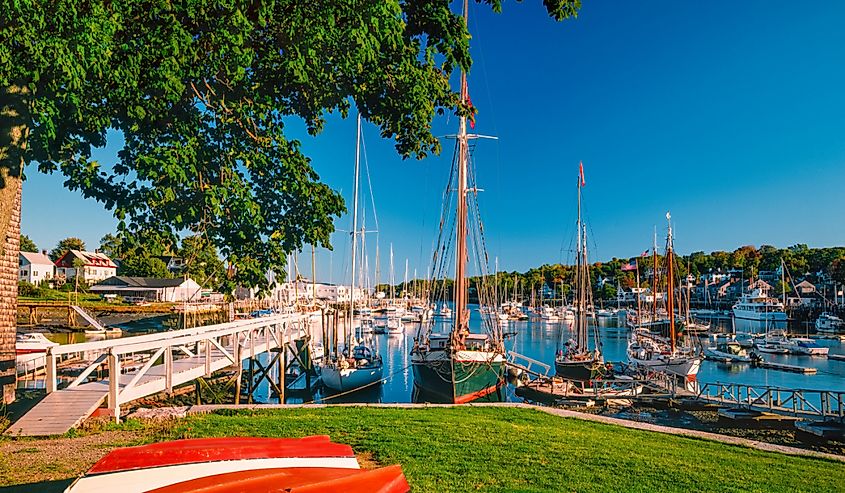 Row boats lay on grass in front of fishing and recreational boats in the harbor of Camden, Maine in the New England