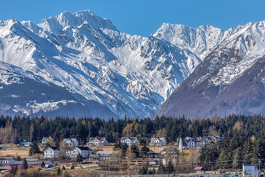 Snowy mountain range under a clear blue sky, with a forested area and a quaint town with white houses nestled at the base, conveying serenity.