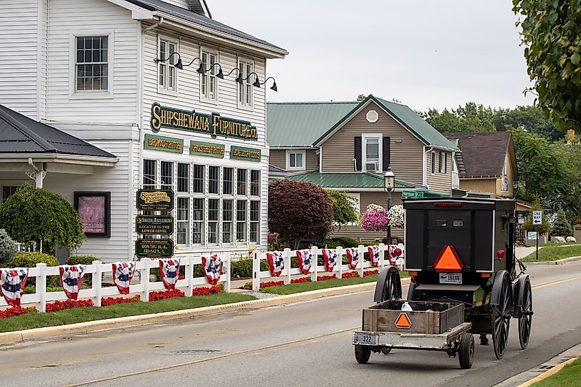 A black Amish horse-drawn buggy towing a small trailer drives along a paved street in Shipshewana, Indiana. On the left, a white building labeled 'Shipshewana Furniture Co.' is decorated with red, white, and blue bunting along a white fence, with neatly landscaped flowers and neighboring houses in the background.