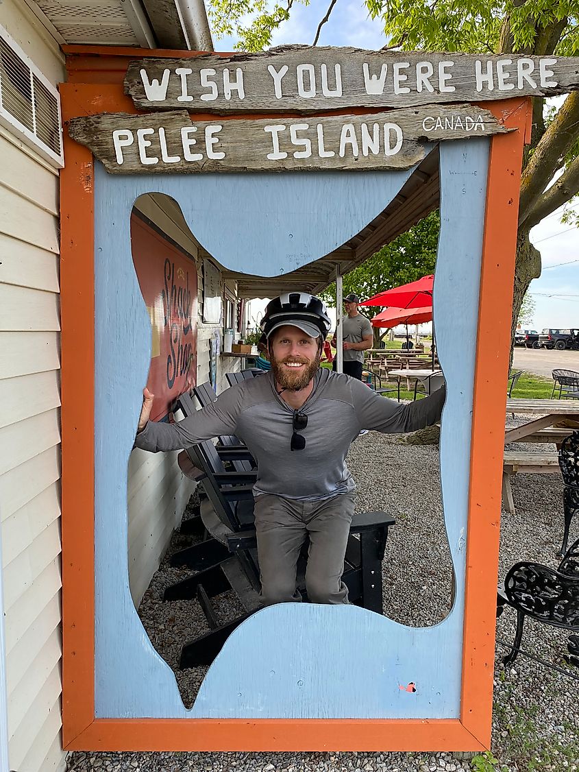 A man with a bike helmet on poses for a picture beneath a wooden sign that says "Wish You Were Here. Pelee Island, Canada."