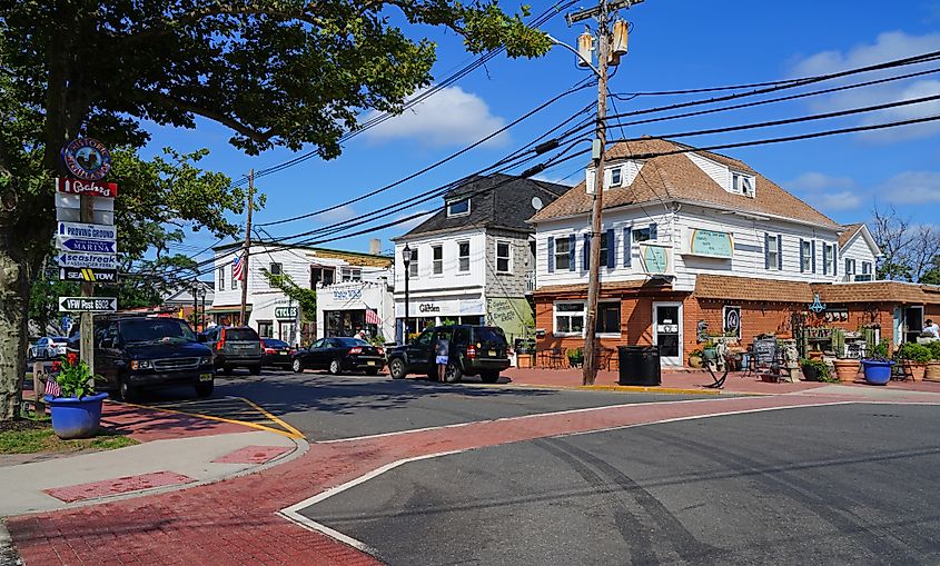 Sunny street-corner view in Highlands, New Jersey, with small shops and restaurants, parked cars, overhead utility wires, and a signpost listing local destinations under a bright blue sky.