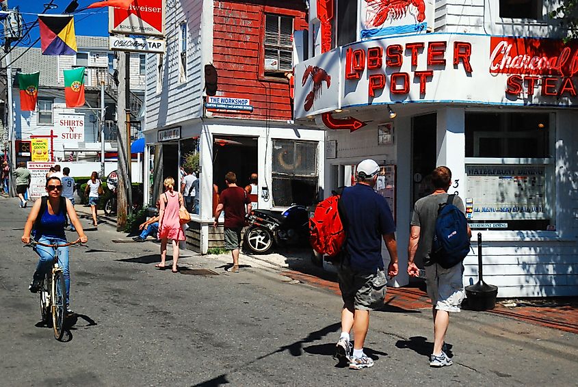 Busy street scene in Provincetown, Massachusetts with pedestrians and a cyclist passing colorful storefronts, including a seafood restaurant with a “Lobster Pot” sign.