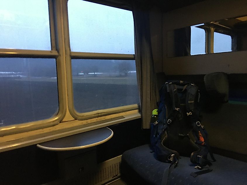 A trekking backpack sits on the bench of a train sleeping booth. A bare countryside can be seen out the window. 