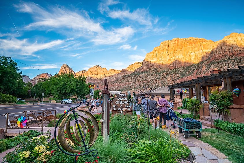 A scenic street in Zion Canyon with vibrant shops, colorful flowers, and people strolling. Red mountains are illuminated by the setting sun under a blue sky.