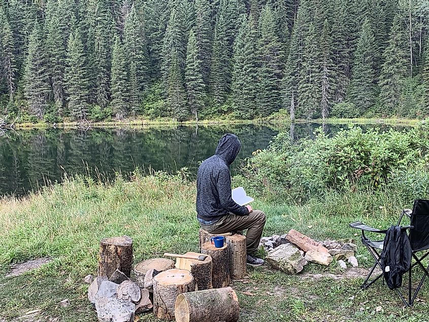 A man in a black hoody reading a book beside a lakeside fire pit. 