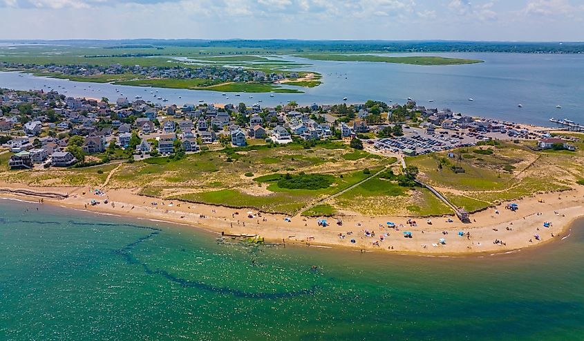 Aerial view of a sandy beach with colorful umbrellas and people relaxing. Behind the beach, a lush green area and residential neighborhood are visible, bordered by calm, blue water under a partly cloudy sky. The scene conveys a serene summer day.