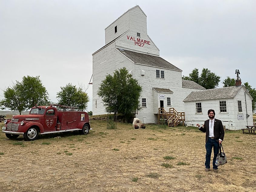 A man in old fashioned attire stands before a historic grain elevator and an antique fire engine.