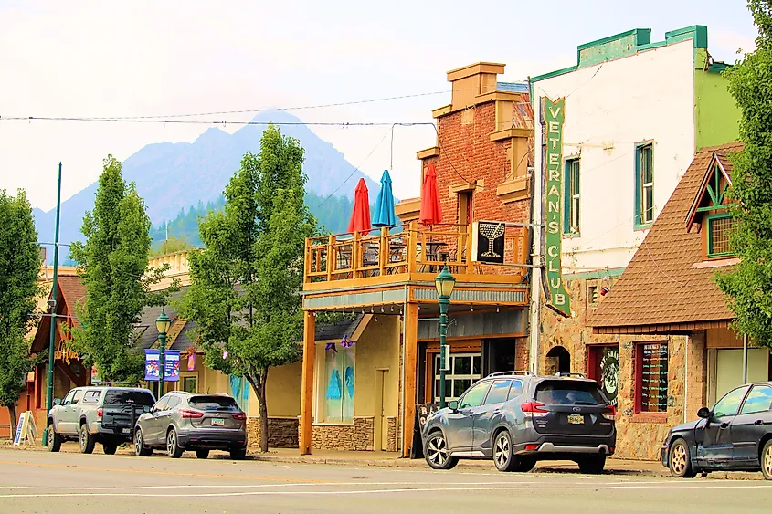 A picturesque small-town street with colorful brick buildings, a Veterans Club sign, outdoor seating with umbrellas, parked cars, and a mountain backdrop.