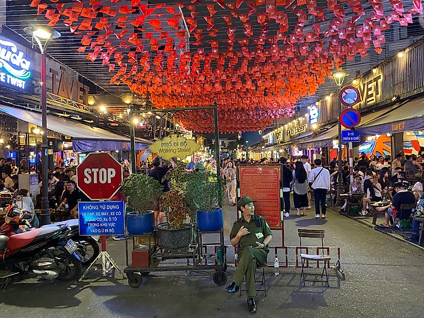 A guard dressed in the typical all green Vietnamese garb sits at the entrance of a busy nightlife street.