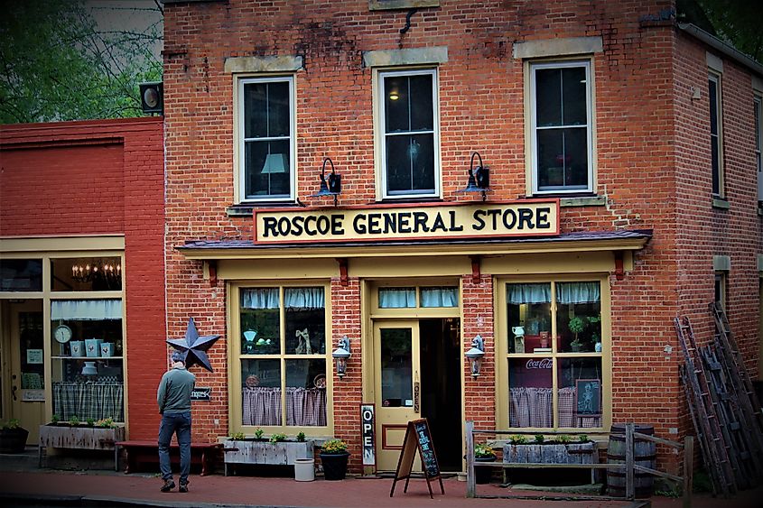 Red-brick storefront with a sign reading "Roscoe General Store," featuring tall second-story windows, display windows with curtains, an open doorway, planters, and a person standing on the sidewalk outside.