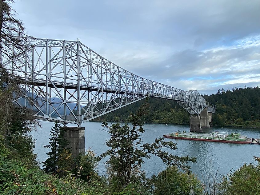 A cargo boat passes beneath a massive steel bridge that spans the lush Columbia River Gorge