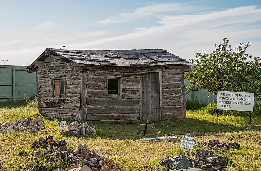 Gray wooden cabin, house, taken from Pine Ridge Indian Tribe and set with display of petrified wooden pieces under light blue cloudscape. Via Shutterstock / Claudine van Massenhove