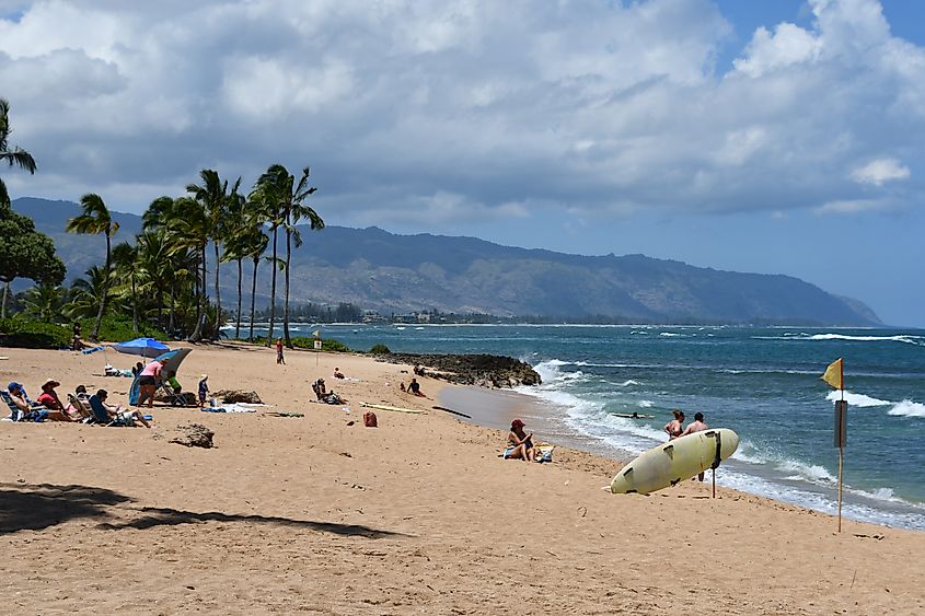 Beachgoers relax and surf at Haleiwa Alii Beach Park on Oahu, Hawaii, with palm trees, rolling waves, and mountains under a partly cloudy sky.