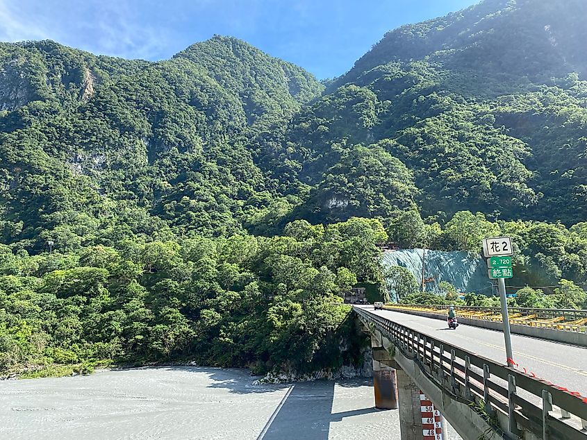 A motorcyclist crossing a river bridge toward steep, lush mountains.