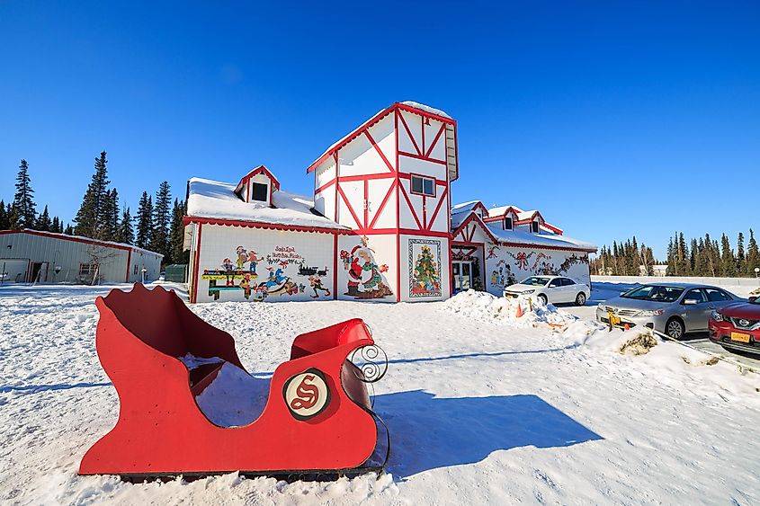 A festive scene with a red sleigh in the foreground and a Christmas-themed building adorned with murals of Santa and elves. Snow covers the ground under a clear blue sky.