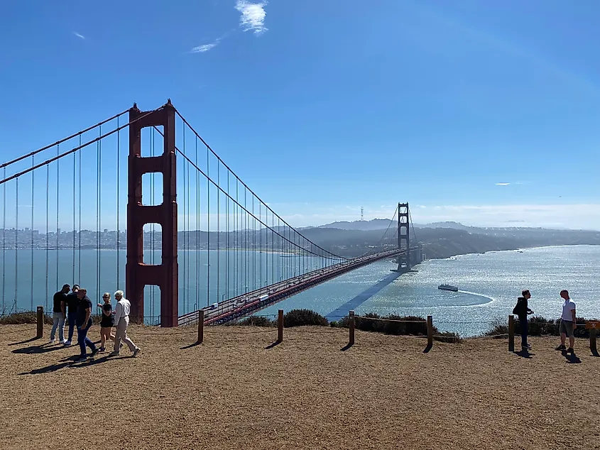 Small groups of people gather on a lookout above the Golden Gate Bridge. Below, a ferry can be seen making a U-turn.