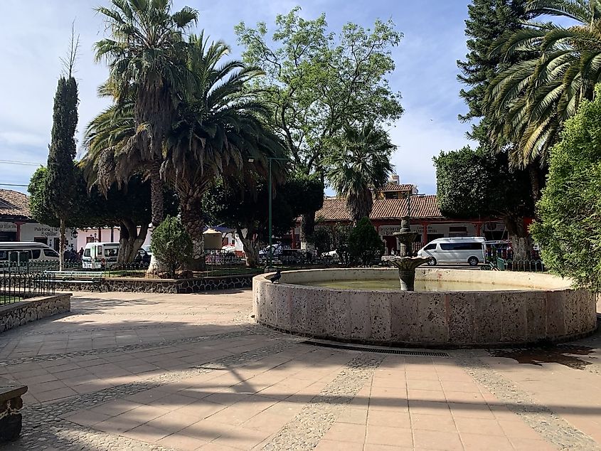 A public fountain in the middle of a quiet town square, with micro-buses or "colectivos" waiting in queue.