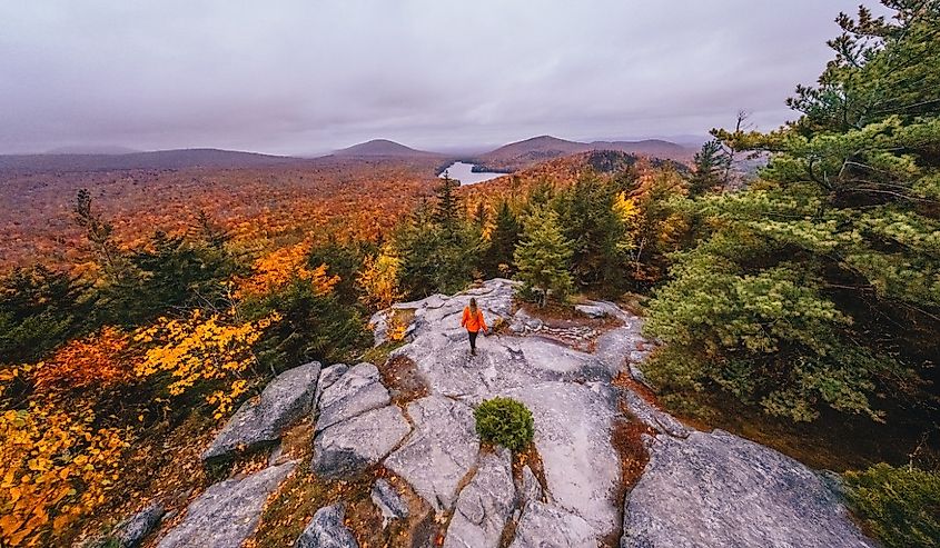 Epic Wide New England Fall Foliage Landscape Aerial Female Hiker in Orange Coat Stands on Owl Head Mountain Vermont Overlooking Colorful Trees Autumn