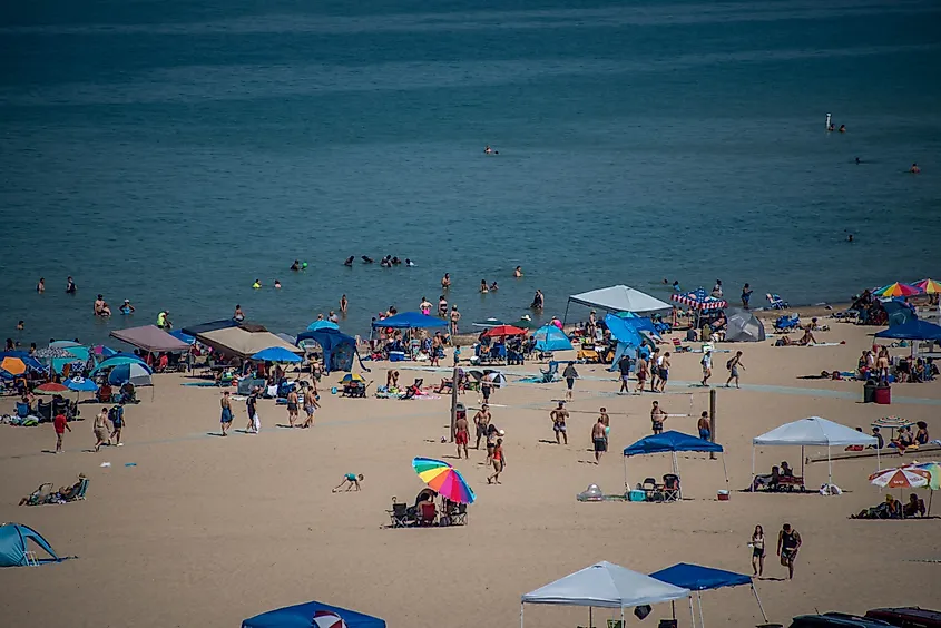A wide sandy beach in Porter, Indiana, is filled with people swimming, relaxing, and playing under colorful umbrellas and pop-up tents. Groups gather near the water's edge while others walk or play games on the sand, with the calm blue waters of Lake Michigan stretching into the background.