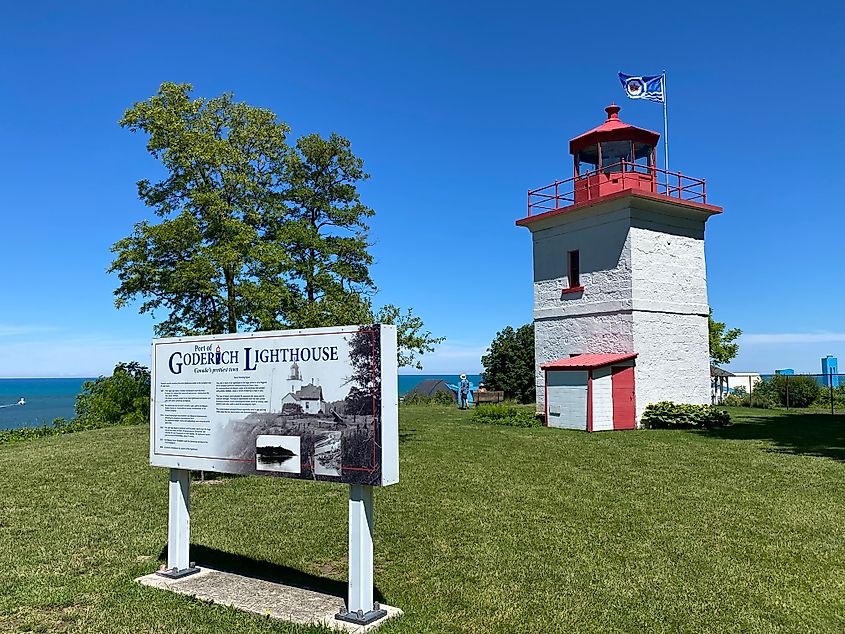 An interpretive sign for the Port of Goderich Lighthouse stands before a short, rectangular white and red lighthouse.