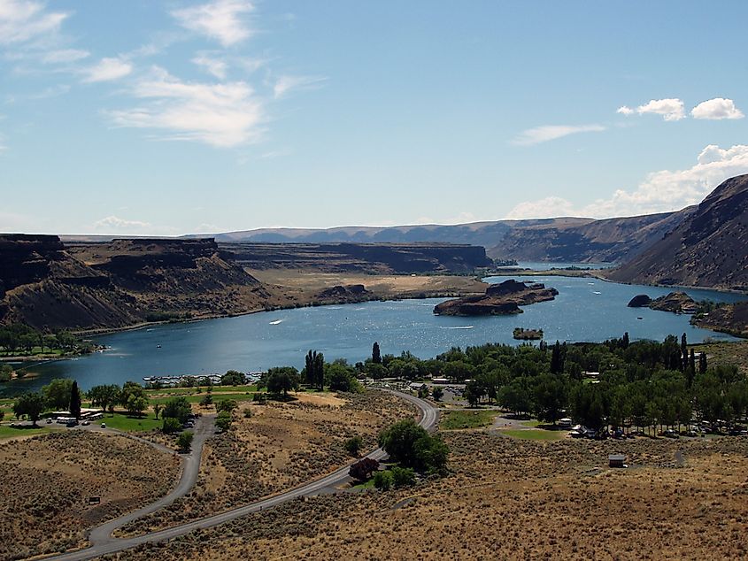 A panoramic view of a town in autumn, with colorful foliage in the foreground, a calm river, and rolling hills under a partly cloudy sky in the background.