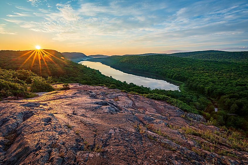 Sunrise casts golden rays across a rocky overlook above Lake of the Clouds in Porcupine Mountains Wilderness State Park, with the winding lake surrounded by dense green forest in Michigan's Upper Peninsula.