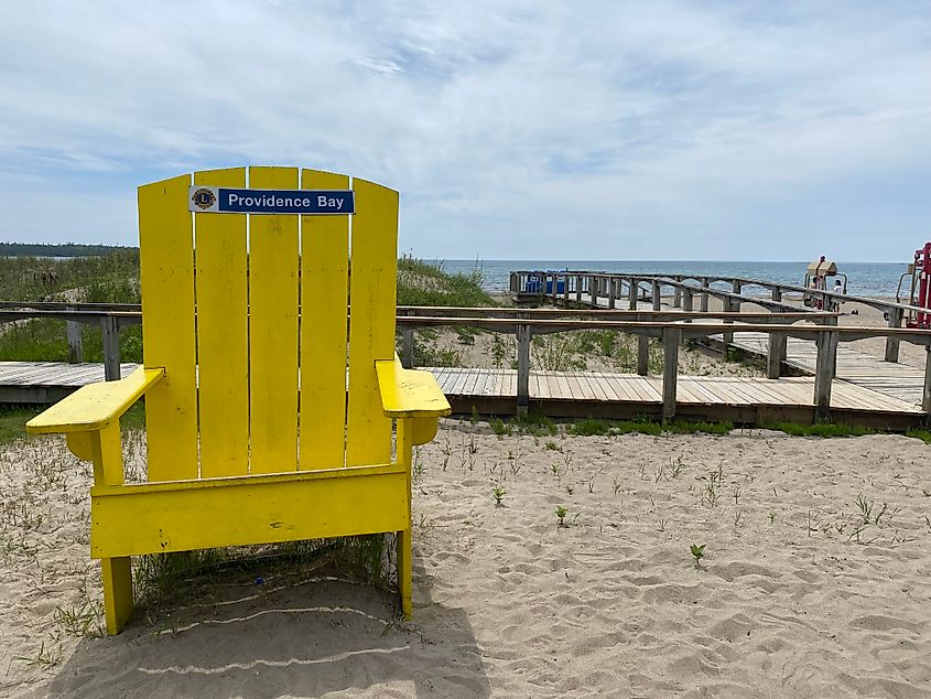 A large yellow cottage chair signed "Providence Bay" stands before a beach, playground, and boardwalk. 