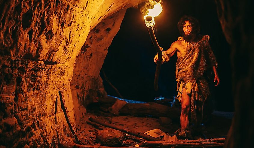 Primeval Caveman Wearing Animal Skin Exploring Cave At Night, Holding Torch