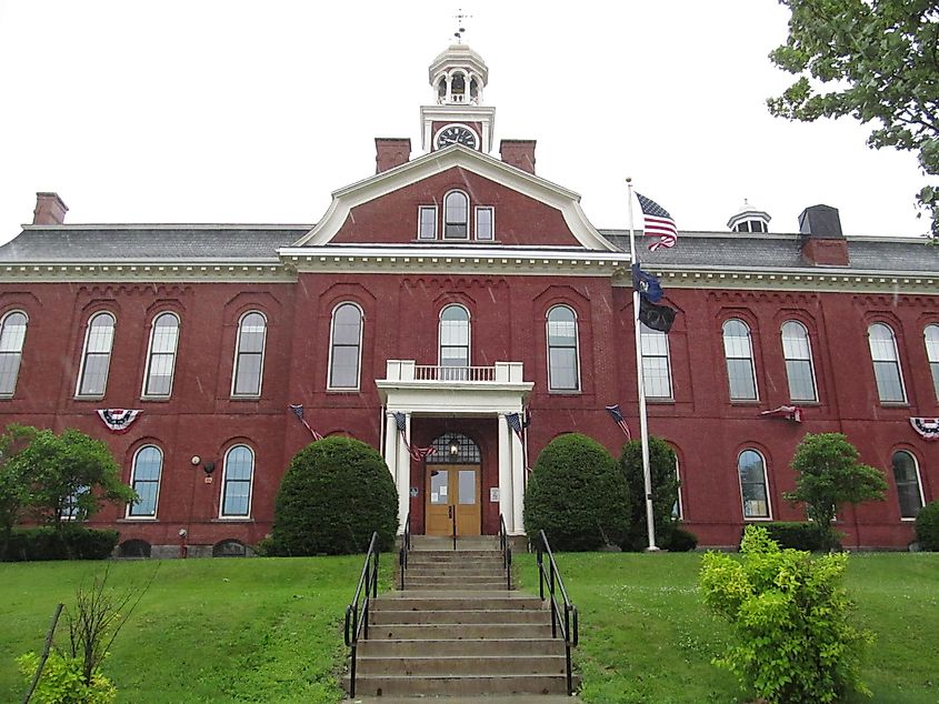 Historic red brick courthouse with arched windows, central entrance, and clock tower. American flag flutters in front, surrounded by lush greenery.