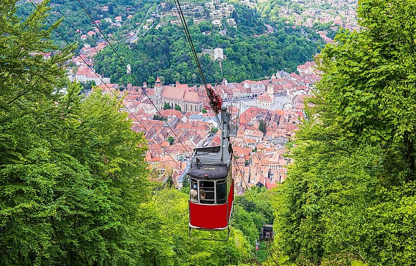 A red cable car rises up a tree-covered mountain above a Romanian old town