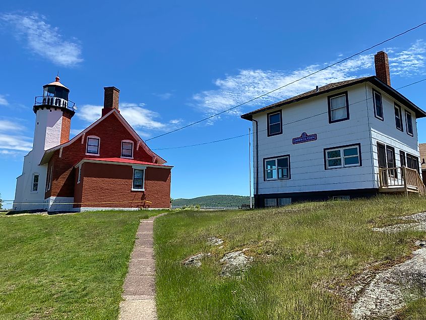 A red-bricked lighthouse stands beside a white-panelled commercial fishing building on a green grassy hill. 