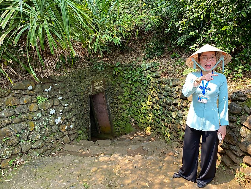 A Vietnamese woman wearing a typical pointed farmer's hat stands outside of a small tunnel entrance.