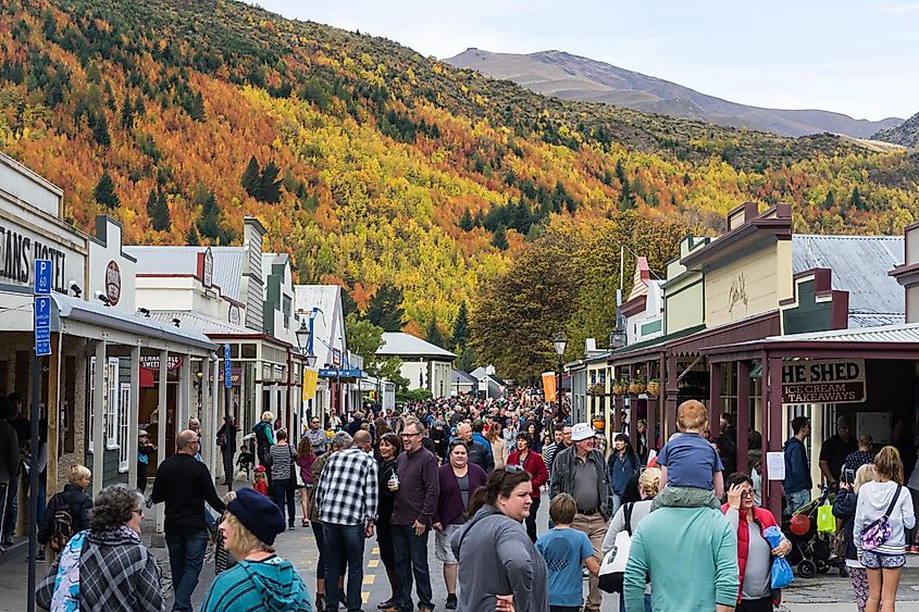 People are walkin along the historic tree walk and talking to each other at the Arrowtown Autumn Festival. Background are some trees and historic buildings.