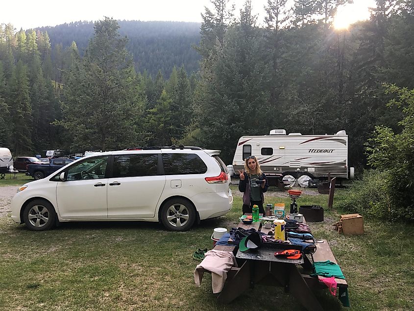 A blond woman giving the peace sign next to a white minivan and a picnic table full off camping goodies.