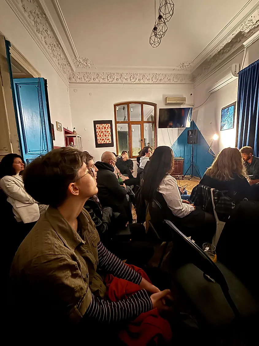 A man sits and reads poetry in front of a small audience in an intimate evening setting.